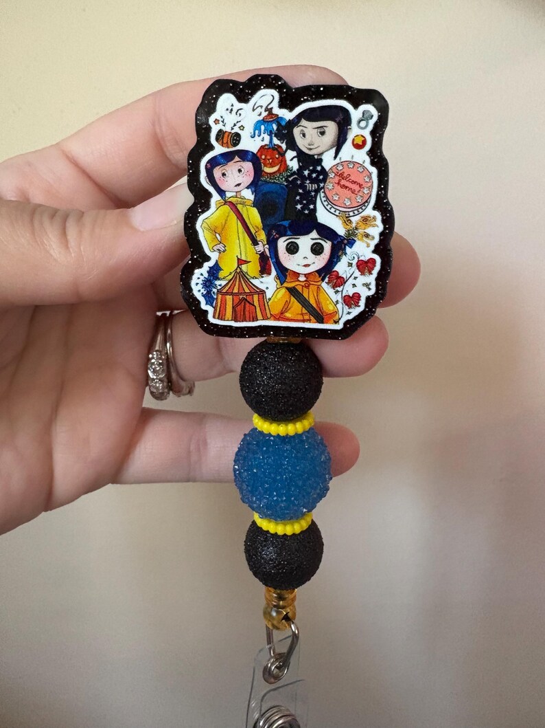 May include: A black and glittery badge reel featuring a colorful illustration of characters from a movie. The badge reel has three decorative beads in black, blue, and yellow. The reel is attached to a clear plastic clip.