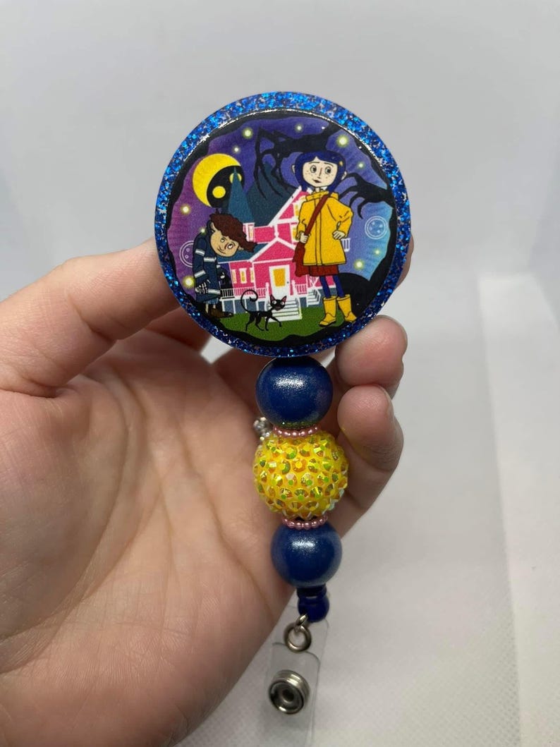 May include: A blue and yellow beaded retractable badge holder with a round, glittery, blue and white button featuring a cartoon image of two children standing in front of a pink house with a black cat. The button has a dark blue border.