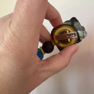 May include: A gold-toned brooch with a circular design, featuring a dark, textured accent and a beaded strand in blue, black, and yellow. The brooch has a pin closure.