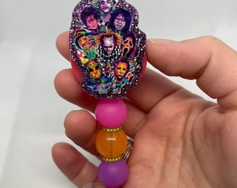 May include: A badge reel featuring a purple glitter-encased topper decorated with a collage of various horror movie characters. Below the topper, the reel includes a vertical arrangement of three smooth, round beads in pink, orange, and purple.