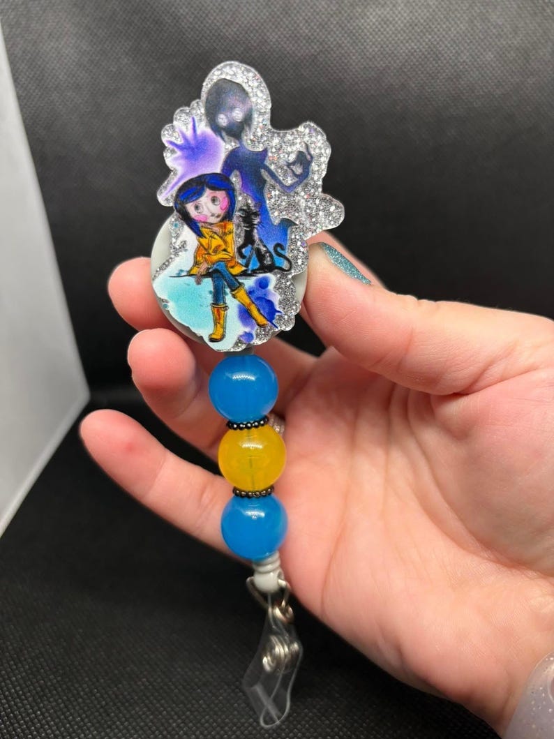 May include: A blue and yellow beaded retractable badge holder with a round, glittery charm featuring a cartoon girl with a black cat and a shadowy figure.