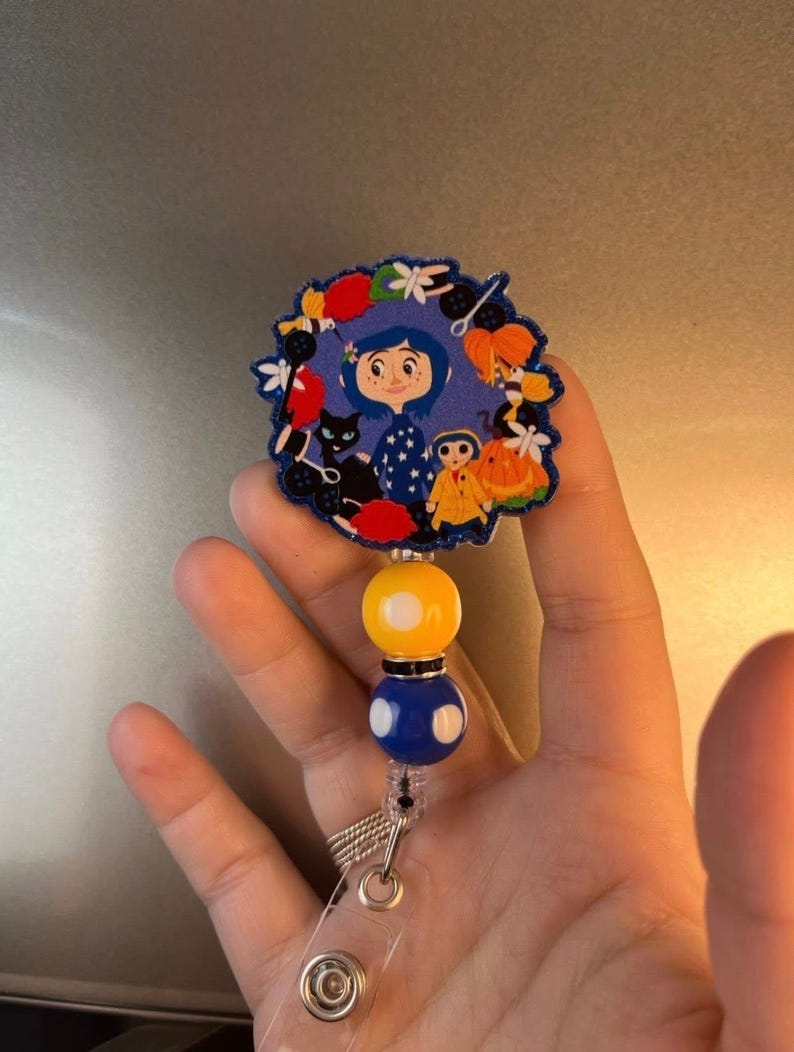 May include: A blue and yellow beaded retractable badge holder with a round, colorful, cartoon character charm featuring a girl with black hair and a black cat.