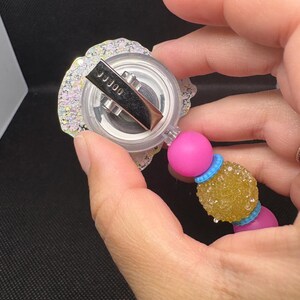 May include: A retractable badge reel with a clear, glittery, floral-shaped top. The reel has a silver clip and is attached to a pink bead, a yellow textured bead, and a blue bead. The text "JJJ00" is visible on the clip.