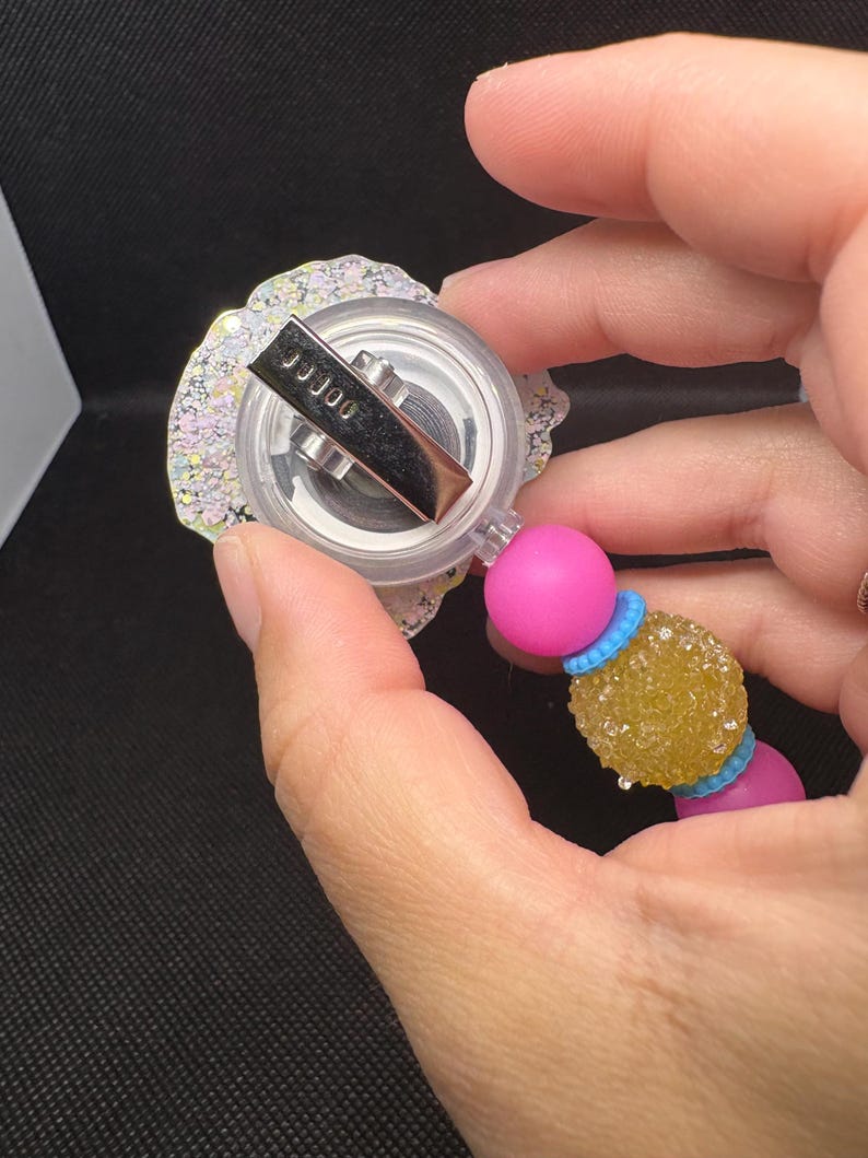 May include: A retractable badge reel with a clear, glittery, floral-shaped top. The reel has a silver clip and is attached to a pink bead, a yellow textured bead, and a blue bead. The text "JJJ00" is visible on the clip.