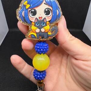May include: A badge reel featuring a cartoon girl with blue hair and a black cat. The badge reel has blue and yellow beads and a gold glitter border. The reel is attached to a metal clip.