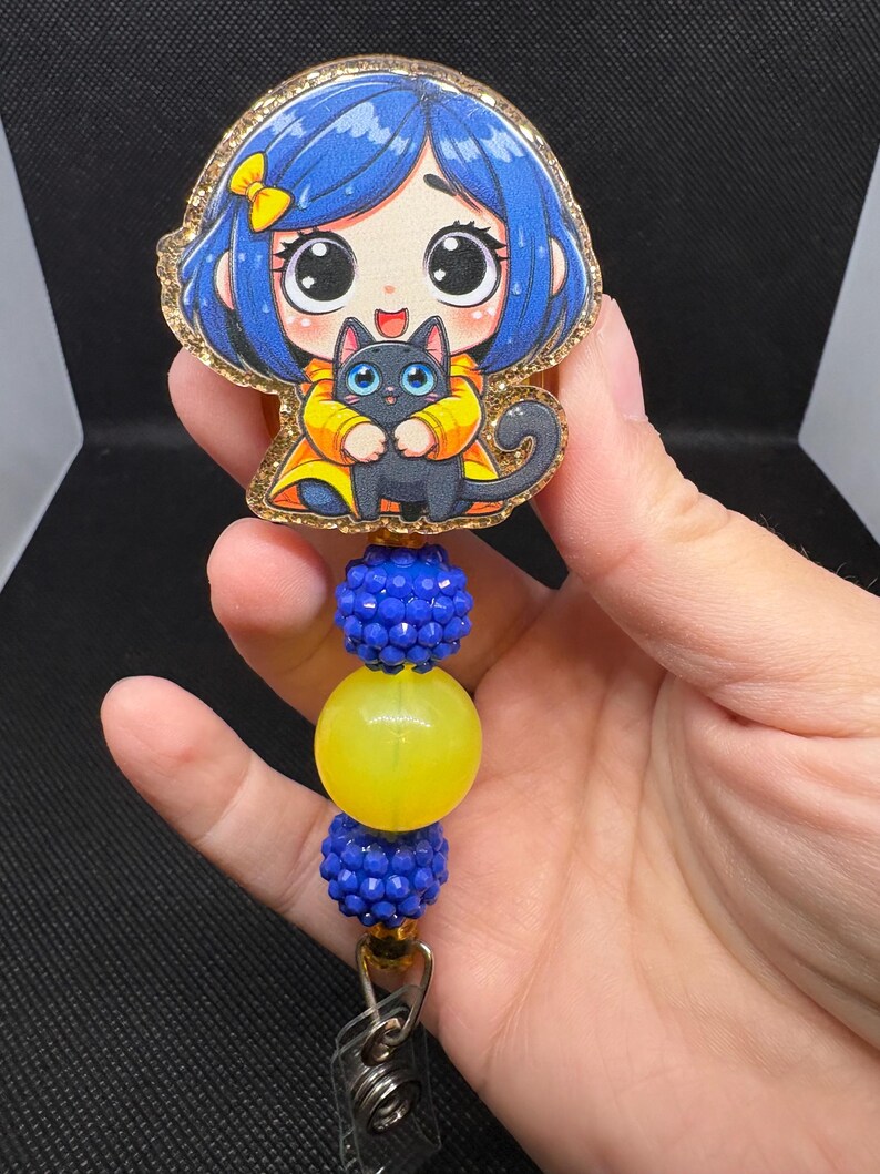 May include: A badge reel featuring a cartoon girl with blue hair and a black cat. The badge reel has blue and yellow beads and a gold glitter border. The reel is attached to a metal clip.