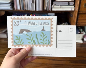 May include: A postcard featuring a stamp design with the text "80¢ CHANNEL ISLANDS." The stamp depicts a seascape with a rock formation, blue water, and green seaweed. The postcard is held against a wooden background.