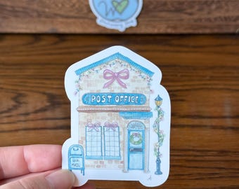 May include: A sticker featuring a whimsical illustration of a post office. The building is light blue with a pink bow, a wreath on the door, and the words "POST OFFICE" above the entrance. A mailbox is on the left side, and a "Wild Hearts" sticker is in the background.