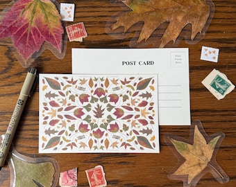May include: A postcard with a symmetrical leaf pattern in red, green, and brown, next to a blank postcard. The scene includes leaf-shaped stickers, vintage stamps, and a Micron 05 pen, all arranged on a wooden surface.