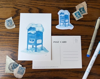 May include: Watercolor illustration of a blue mailbox covered in snow, with a matching sticker and postcard. Vintage stamps and pens are arranged around the artwork on a wooden surface. The postcard reads "POST CARD".