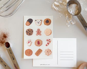 May include: A postcard with a watercolor illustration of various Christmas cookies, including gingerbread men, candy canes, and a snowman. The postcard has a blank space for writing a message and the text "@Wildhearts.Art" at the bottom.