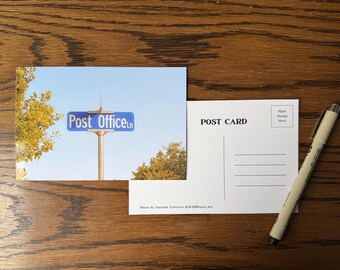 May include: A rectangular postcard featuring a blue street sign that reads Post Office Ln against a clear sky with yellow autumn trees. The reverse side is white with the text POST CARD, a stamp box, address lines, and Photo by Autumn Universe @WildHearts.Art.