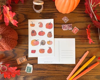 May include: A postcard with a watercolor illustration of pumpkins in various shades of orange and red. The postcard has a blank space for writing a message and an address on the back. The postcard is on a wooden surface with fall leaves and other decorative items.