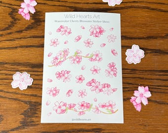 May include: A sticker sheet with watercolor cherry blossom designs and loose stickers scattered around it. The sheet reads "Wild Hearts Art Watercolor Cherry Blossoms Sticker Sheet" with the artist's handle. The stickers are pink.