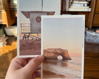 May include: Two postcards featuring coastal scenes. One shows a lifeguard tower, the other a natural rock arch over the ocean. Text on the cards reads "Natural Bridges State Beach, Santa Cruz, California."