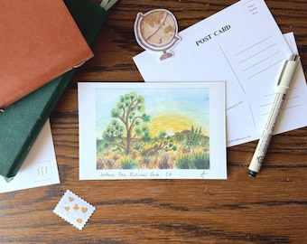 May include: A watercolor postcard featuring a Joshua Tree landscape with a sunset. The postcard is on a wooden surface with a pen, postcards, a globe sticker, and a stamp. The text "Joshua Tree National Park, CA" is visible.
