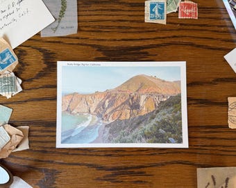 May include: A postcard featuring Bixby Bridge in Big Sur, California, with a mountain and ocean view. The postcard is surrounded by vintage stamps, a handwritten note, and other ephemera on a wooden surface.