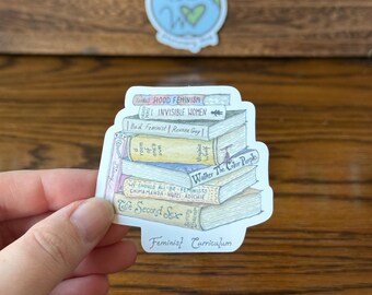 May include: A sticker featuring a stack of books with titles like "Hood Feminism" and "Invisible Women." The sticker also includes the text "Feminist Curriculum" and a small globe graphic. The sticker is held by a person.