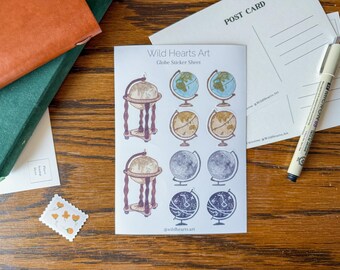 May include: A sheet of globe stickers from Wild Hearts Art, featuring various globe designs in blue, tan, and gray. The stickers are on a white background, with a postcard and pen nearby. A stamp and books are also visible.