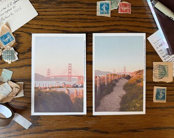 May include: Two postcards featuring the Golden Gate Bridge in San Francisco, California. The postcards are on a wooden surface with stamps, receipts, and a roll of tape. The bridge is a vibrant red against a blue sky.