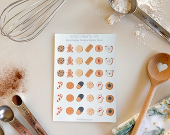 May include: A sheet of mini holiday cookie stickers with various designs, including gingerbread men, bears, and candy canes. The sheet is surrounded by baking tools like a whisk, measuring spoons, and a wooden spoon, suggesting a baking theme.