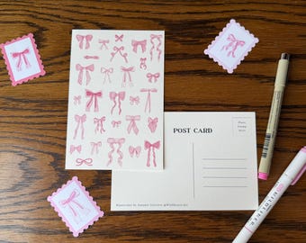 May include: A collection of stationery items on a wooden surface. Includes a postcard, stickers, and a sheet of paper with pink bow illustrations. Two pens are also present. The postcard reads "POST CARD". The stickers and paper feature watercolor designs.