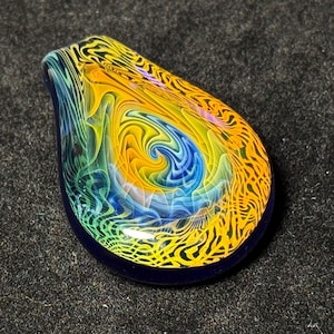 Könnte beinhalten: Ein tropfenförmiger Glasanhänger mit einem wirbelnden Design. Der Anhänger hat eine zentrale blaue Spirale, umgeben von gelben, grünen und orangen Mustern. Die Ränder sind tiefblau und erzeugen einen auffälligen Kontrast.