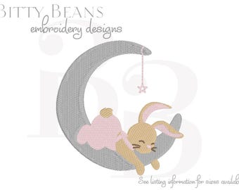May include: An embroidered design featuring a sleeping bunny on a gray crescent moon. The bunny is tan with pink accents, and a pink star hangs from the moon. The text "Bitty Beans embroidery designs" is at the top.