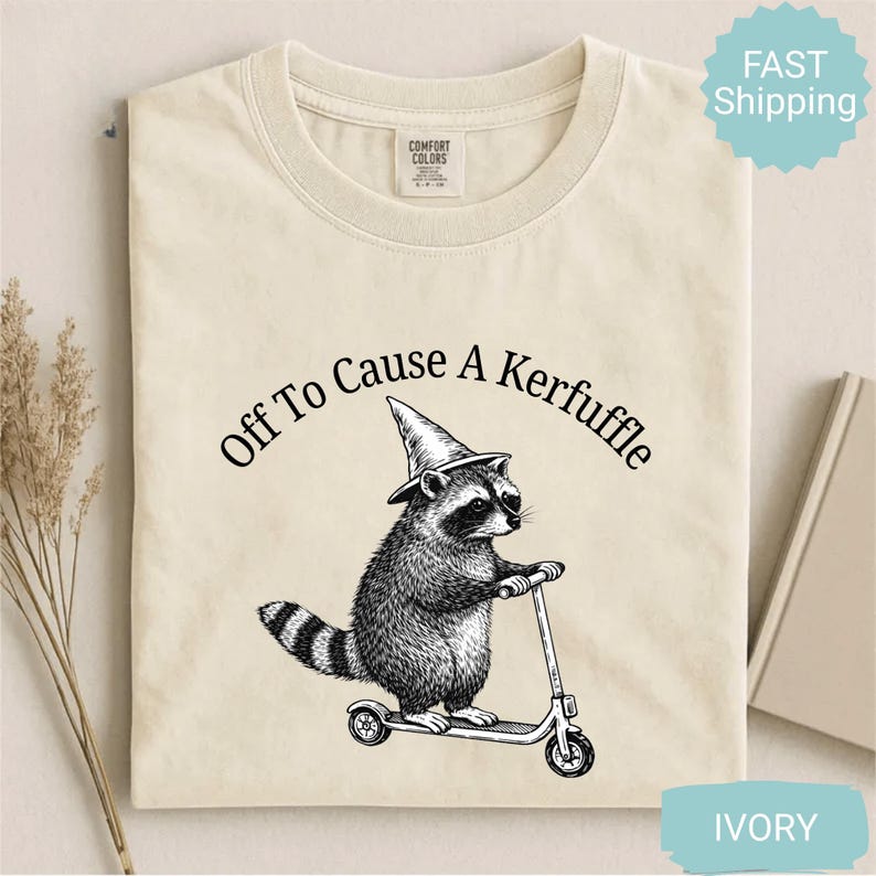 May include: Ivory-colored t-shirt with a black graphic of a raccoon wearing a witch hat and riding a scooter. The text "Off To Cause A Kerfuffle" is arched above the raccoon. The shirt has a crew neck and a tag that says "Comfort Colors".