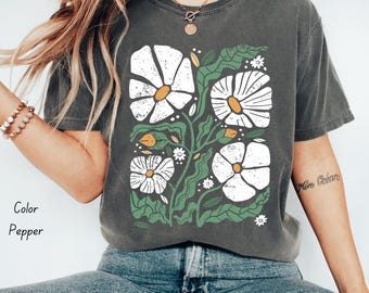 May include: Pepper-colored t-shirt featuring a floral design with white flowers and green leaves. The graphic has a vintage aesthetic. The shirt is a relaxed fit and made of soft material. The text "Color Pepper" is visible on the left side of the image.