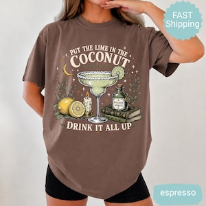 May include: A brown t-shirt featuring a graphic print of a margarita glass, a lime, a candle, a bottle labeled Witches Spirits, and two books. The text reads, Put the lime in the coconut, drink it all up. The design includes stars and a crescent moon.