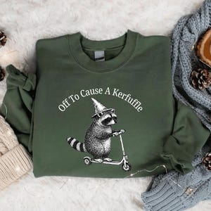 May include: Green sweatshirt with the text "Off To Cause A Kerfuffle" in a curved arc. A raccoon wearing a witch's hat is riding a scooter. The raccoon is black and white, and the scooter is silver.