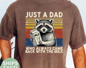 Pode incluir: Camiseta marrom com um guaxinim segurando uma caixa de leite e um copo de leite. O texto diz "JUST A DAD WHO ALWAYS CAME BACK WITH THE MILK". O design tem um estilo retrô e uma aparência vintage.