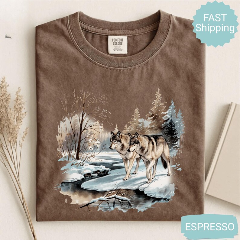 Op de afbeelding: Espresso-kleurig T-shirt met een winterse sc&egrave;ne met twee wolven bij een besneeuwde rivier. De afbeelding bevat bomen en een besneeuwd landschap. Het shirt is gelabeld met "Comfort Colors".