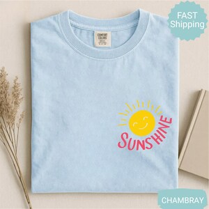 May include: Pale blue t-shirt featuring a yellow smiling sun graphic and the word "Sunshine" in pink. The shirt has a crew neck and a small tag on the upper left. The words "Fast Shipping" are in a turquoise circle.