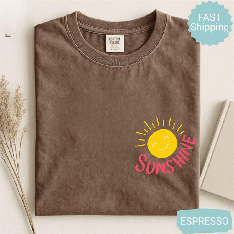 May include: Espresso-coloured t-shirt featuring a yellow smiling sun graphic and the word "Sunshine" in pink. The shirt is folded and has a Comfort Colors tag.
