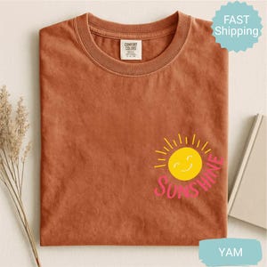 May include: A terracotta-coloured t-shirt with a crew neck. The shirt features a yellow smiling sun graphic with the word "Sunshine" in pink. The shirt is made of a soft material.