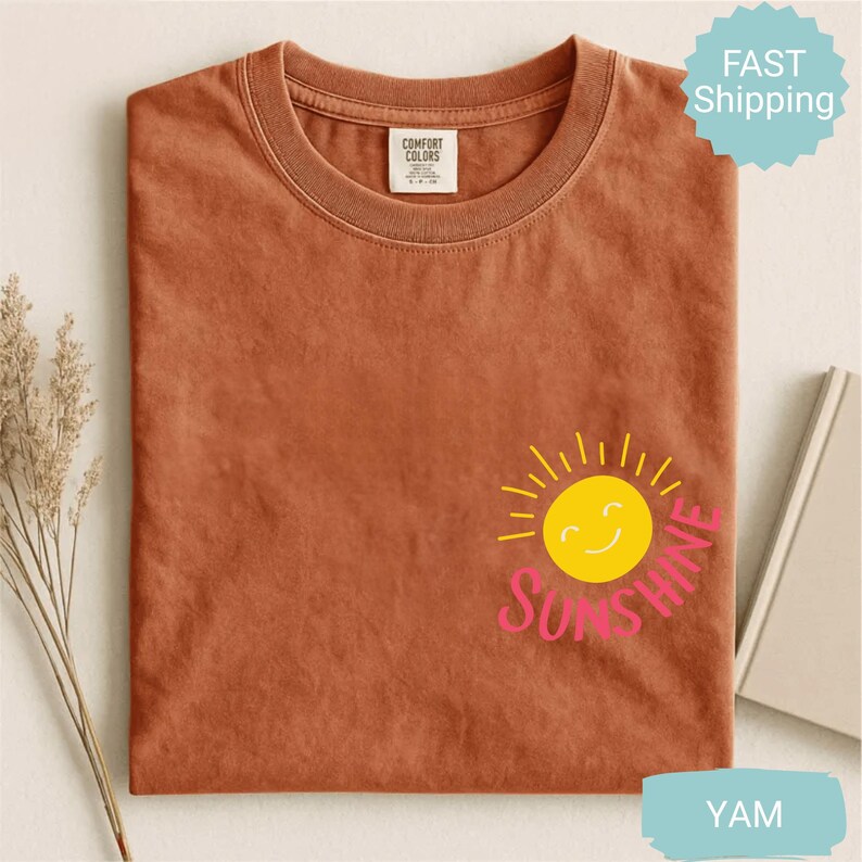 May include: A terracotta-coloured t-shirt with a crew neck. The shirt features a yellow smiling sun graphic with the word "Sunshine" in pink. The shirt is made of a soft material.