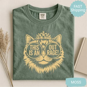 May include: Moss green t-shirt featuring a cat wearing sunglasses and a crown. The sunglasses have the words "THIS IS AN" on the left lens and "OUT RAGE!" on the right lens. The shirt is made of a soft material.