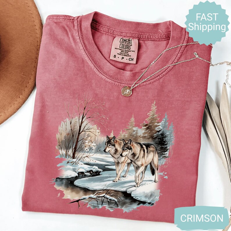 Op de afbeelding: Kleurrijk T-shirt met een winterse sc&egrave;ne van twee wolven bij een besneeuwde rivier. Het shirt heeft een ronde hals en korte mouwen. Een zilveren ketting met een hanger is over het shirt gedrapeerd. Het label luidt "Comfort Colors".