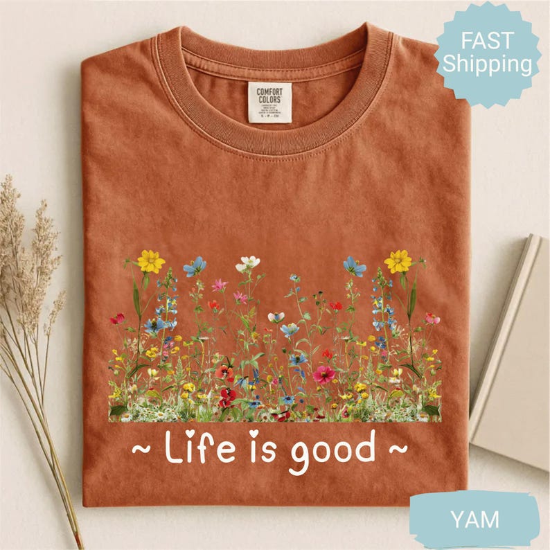 May include: A terracotta-coloured t-shirt with a floral design featuring yellow, blue, pink, and white wildflowers. The text "~ Life is good ~" is printed below the flowers. The shirt has a crew neck and short sleeves.