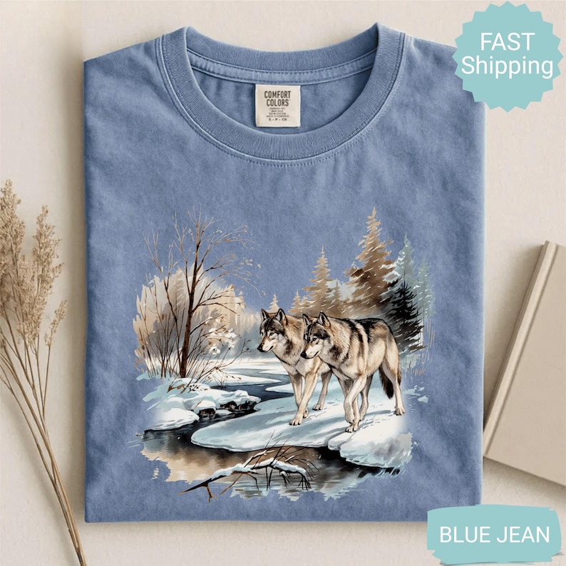 Op de afbeelding: Blauw spijkerbroekkleurig T-shirt met een winterse sc&egrave;ne van twee wolven bij een besneeuwde rivier. De afbeelding bevat bomen en een besneeuwd landschap. Het shirt heeft een ronde hals en een Comfort Colors-label.