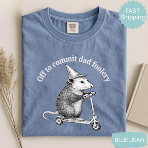 Op de afbeelding: Blauw Comfort Colors T-shirt met een opossum met een tovenaars hoed op een scooter. De tekst "Off to commit dad foolery" staat boven de opossum. Een grappig item.