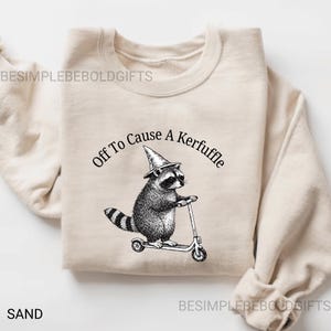 May include: Sand-colored sweatshirt featuring a black and white illustration of a raccoon wearing a witch hat and riding a scooter. The text "Off To Cause A Kerfuffle" is arched above the raccoon. The word "SAND" is printed at the bottom left.