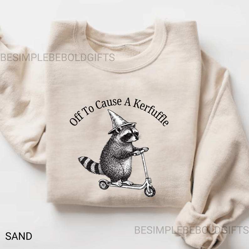 May include: Sand-colored sweatshirt featuring a black and white illustration of a raccoon wearing a witch hat and riding a scooter. The text "Off To Cause A Kerfuffle" is arched above the raccoon. The word "SAND" is printed at the bottom left.