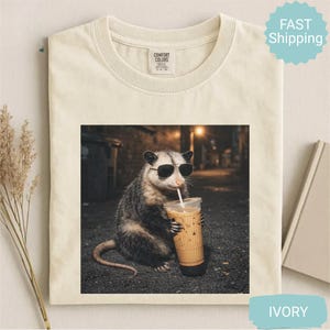 May include: Ivory-colored t-shirt featuring a graphic of an opossum wearing sunglasses and drinking an iced coffee. The opossum is in a dark alley setting. The shirt has a Comfort Colors tag.