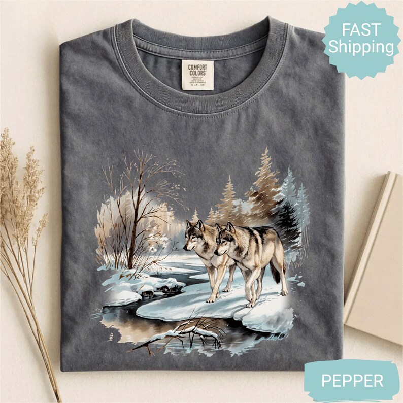 Op de afbeelding: Donkergrijs Comfort Colors T-shirt met een winterse sc&egrave;ne van twee wolven bij een besneeuwde beek. De afbeelding bevat bomen en een besneeuwd landschap. Het shirt heeft een ronde hals en een Comfort Colors label.