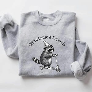 May include: A light gray sweatshirt featuring a raccoon wearing a witch's hat and riding a scooter. The text "Off To Cause A Kerfuffle" is arched above the raccoon. The sleeves are cuffed.