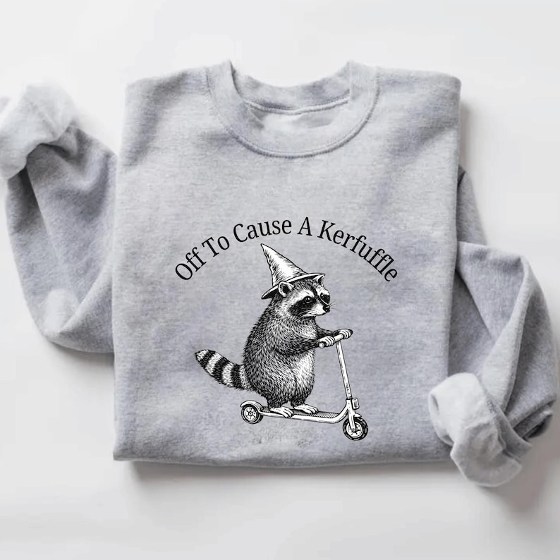 May include: A light gray sweatshirt featuring a raccoon wearing a witch's hat and riding a scooter. The text "Off To Cause A Kerfuffle" is arched above the raccoon. The sleeves are cuffed.