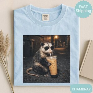 May include: Light blue t-shirt featuring a graphic of an opossum wearing sunglasses and drinking an iced coffee. The opossum is set against a blurred urban background. The shirt has a Comfort Colors tag.